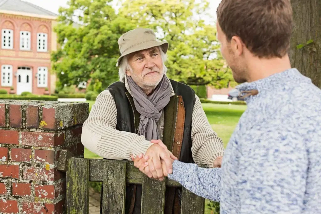 Showing Senior man and mid adult man shaking hands over gate.